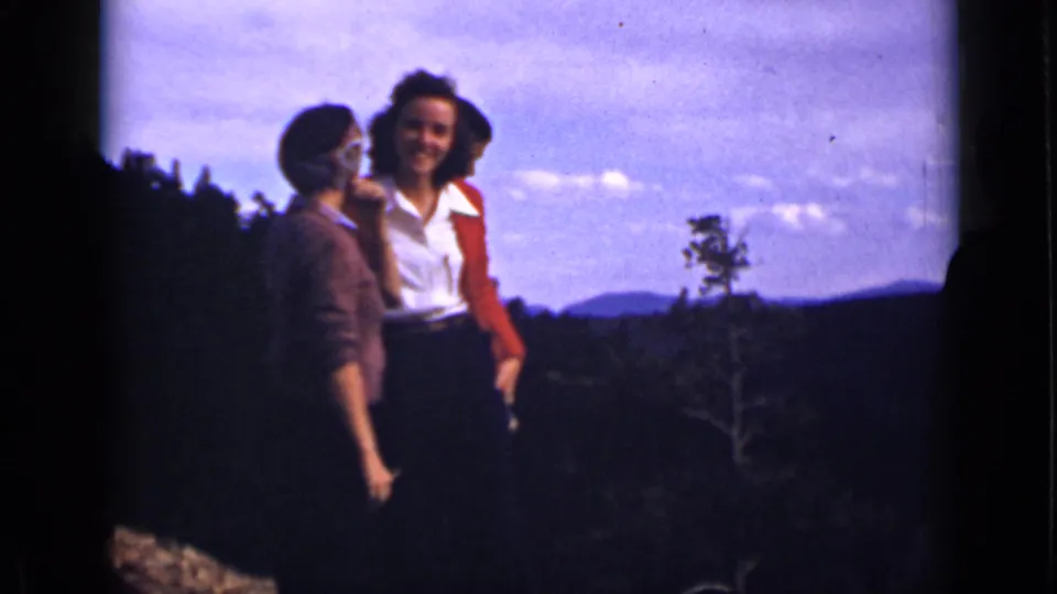 Three women in 1939 Colorado home-movie footage pose on a scenic overlook with wooded slopes, distant mountains, and open sky behind them.
