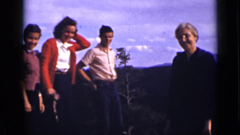 Visitors at a Colorado overlook face the camera, with cardigans, a light shirt, wooded hills, and distant mountains visible in the frame.