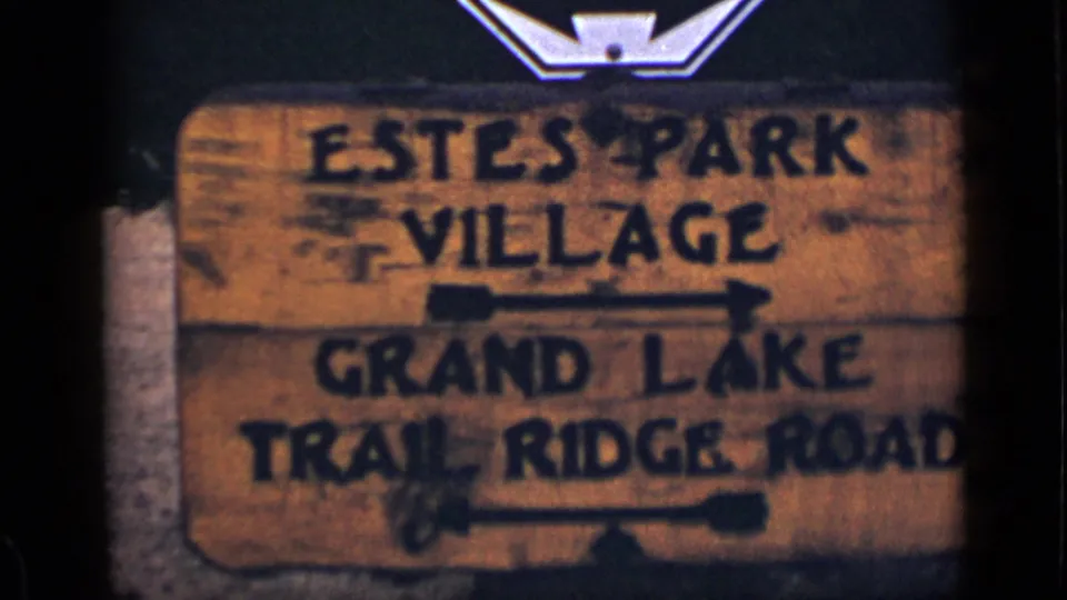 1939 Colorado home-movie frame with a weathered wooden sign for Estes Park Village and Grand Lake Trail Ridge Road.