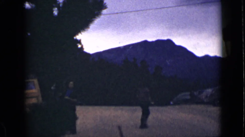 1939 Colorado home-movie frame with a man walking before a dark mountain ridge and roadside trees at dusk or low exposure.