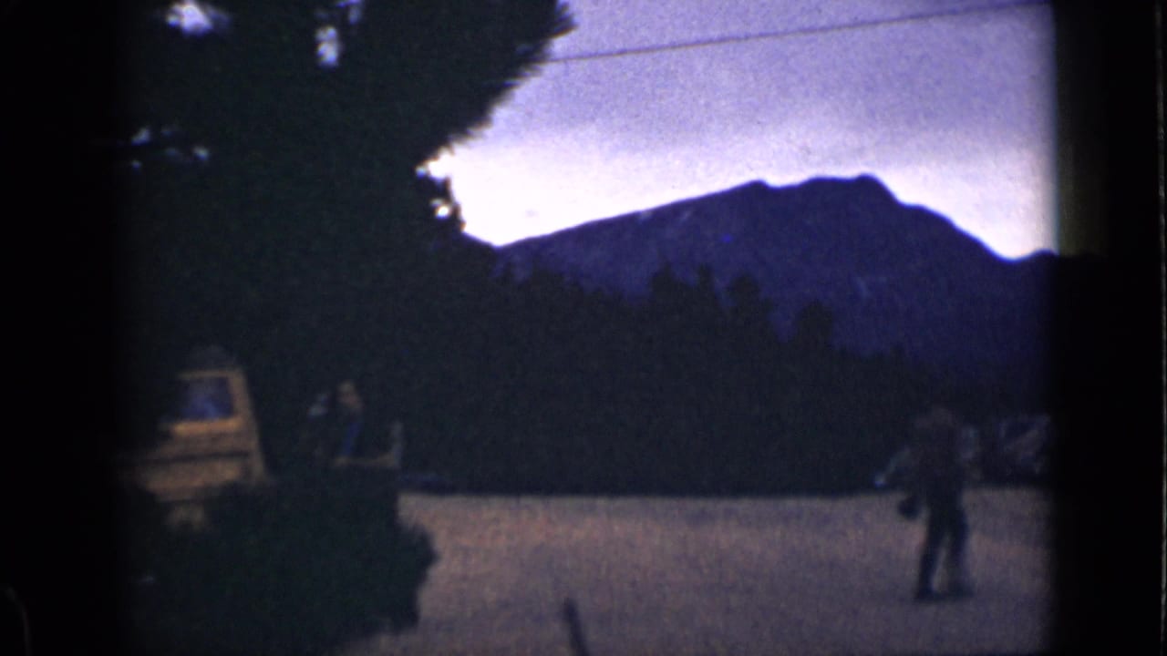 Two adults near a yellow vehicle in a mountain roadside area at dusk or shade.