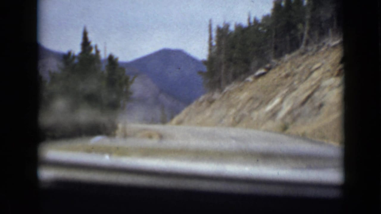 Mountain road seen from inside a moving car, with evergreens, rocky slope, distant peaks, and film-frame borders.