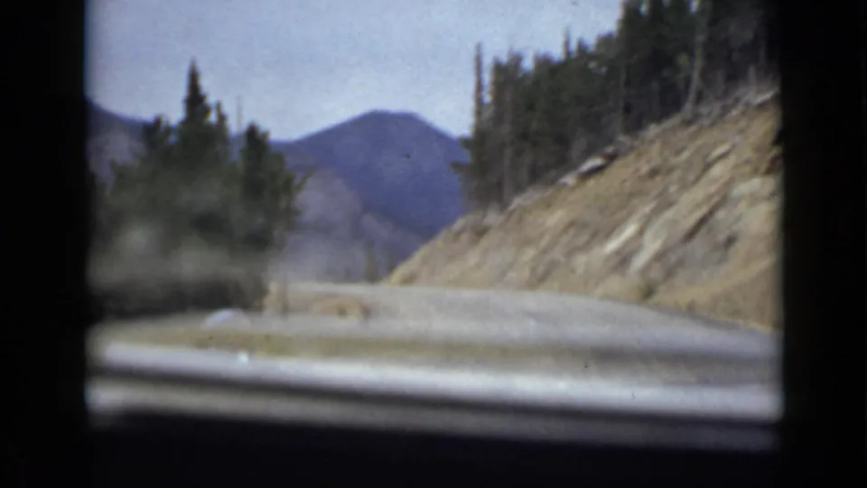 1939 Colorado home-movie view from a car on a paved mountain road, with evergreens, a rocky roadside cut, distant hills, and soft 8mm color.