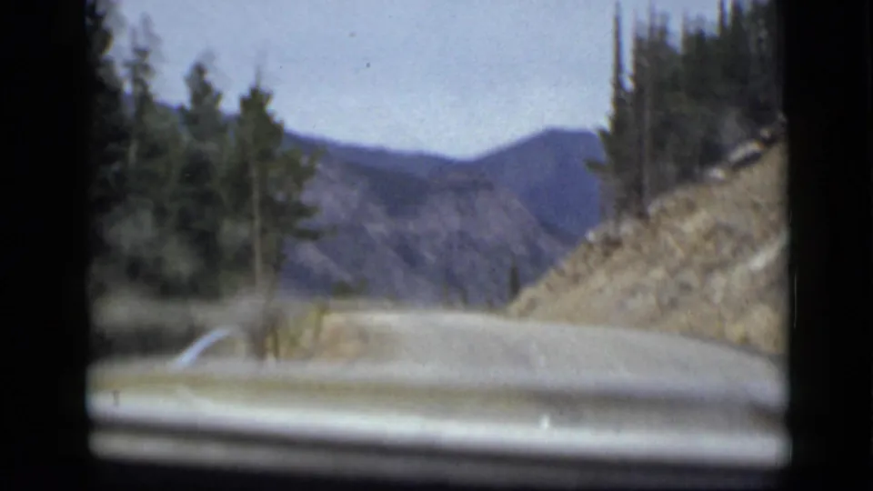 Colorado mountain drive viewed through an 8mm home-movie frame, showing a paved bend, rocky slope, evergreens, and distant ridgelines.