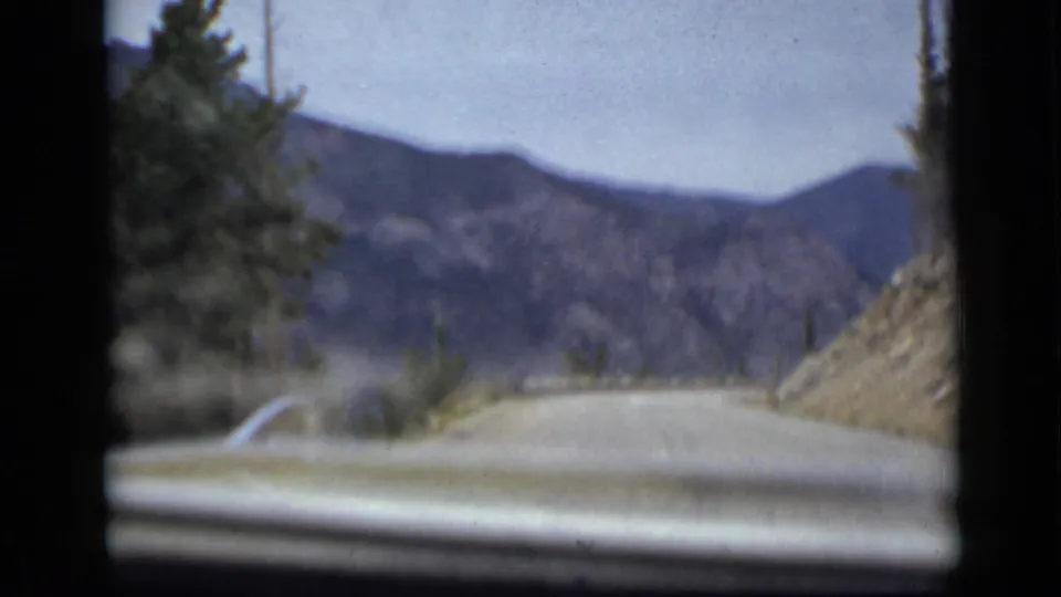 Passenger-side view during a 1939 Colorado mountain road trip, with the car hood low in frame and layered hills beyond the roadway.