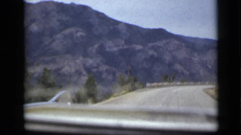 Late-1930s Colorado car-travel view of a paved road curving through rugged mountains, with scattered evergreens and visible 8mm frame edges.