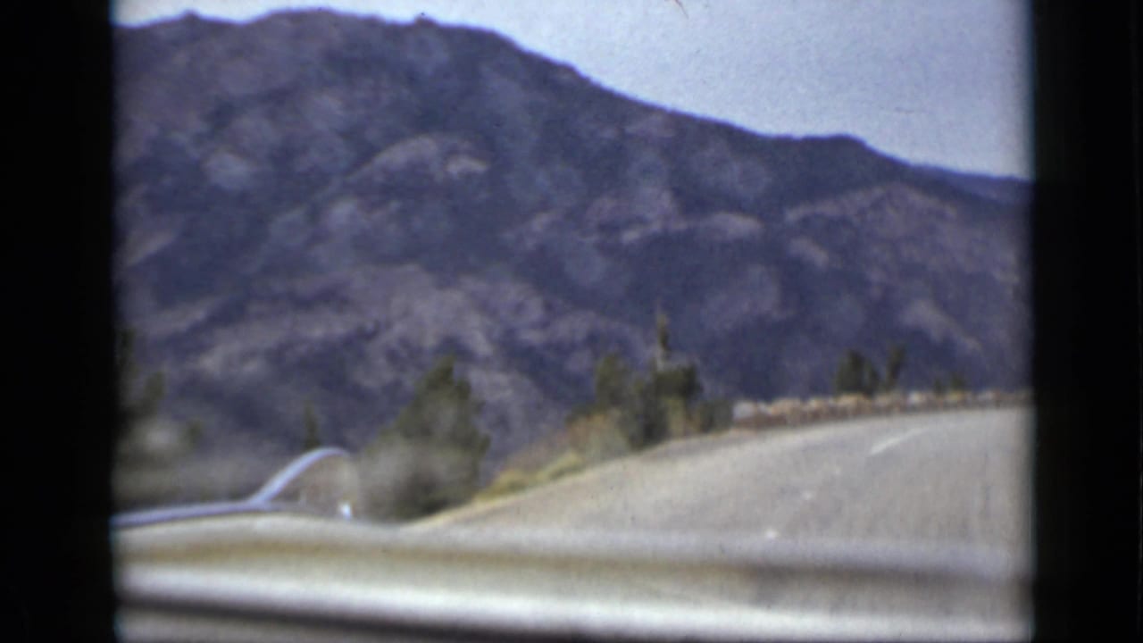 1939 Colorado home-movie frame from inside a moving car, showing a paved mountain road, car hood, stone barrier, scrub, and dark rocky slopes.