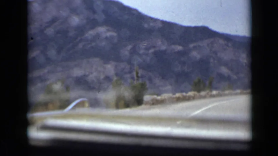 In-car 1939 Colorado road view with motion blur, a low stone barrier, sparse vegetation, and steep rocky mountains beyond the windshield.