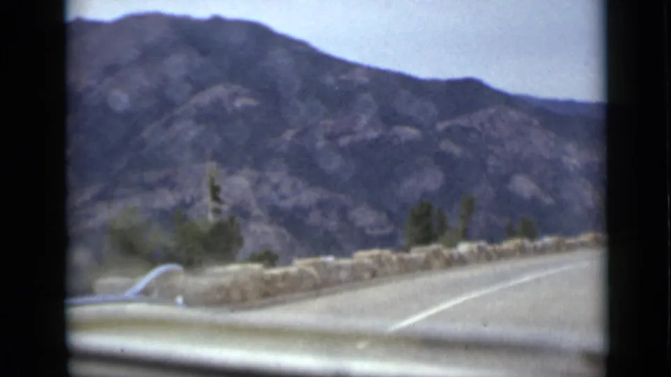 Colorado mountain-road frame from a moving 8mm home movie, showing the car hood, stone guard wall, paved lane, brush, and rocky slopes.