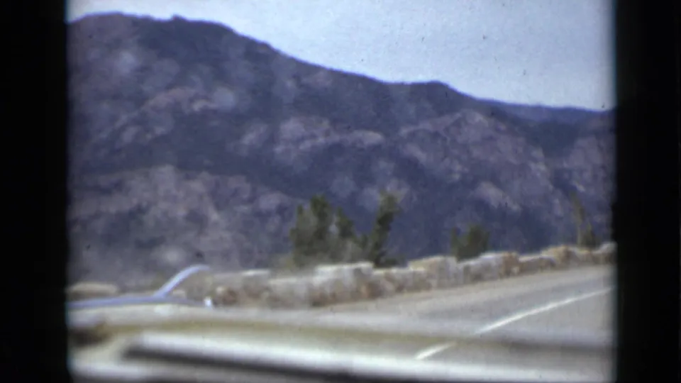 Final 1939 Colorado in-car frame showing a paved mountain road, pale car hood, stone roadside wall, scrub vegetation, and broad rocky hills.