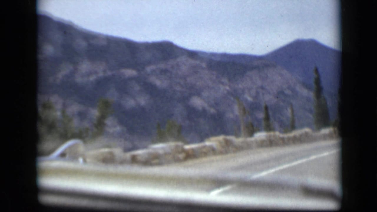 Mountain road seen through a car windshield with rocky slopes and evergreens.