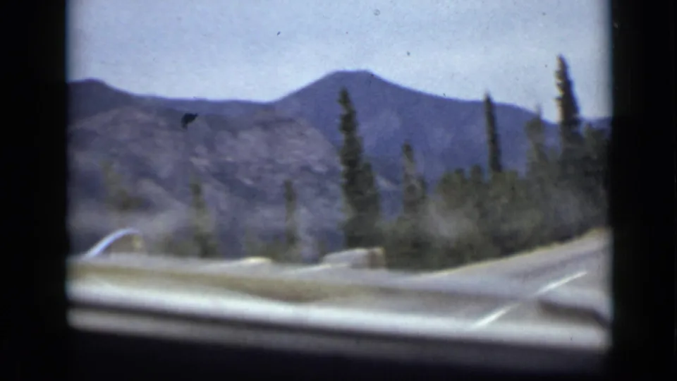 Passenger-view 8mm frame of a Colorado mountain road, with a low windshield edge, roadside stonework, evergreen trees, and distant ridges.