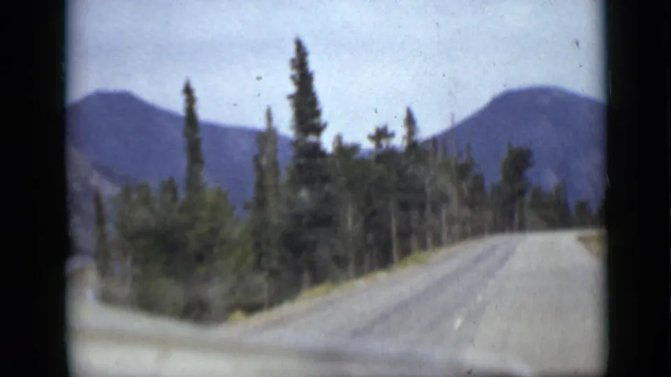 Colorado road-travel frame showing evergreens close to the roadside, blue mountain forms, and a pale sky in soft vintage 8mm color.