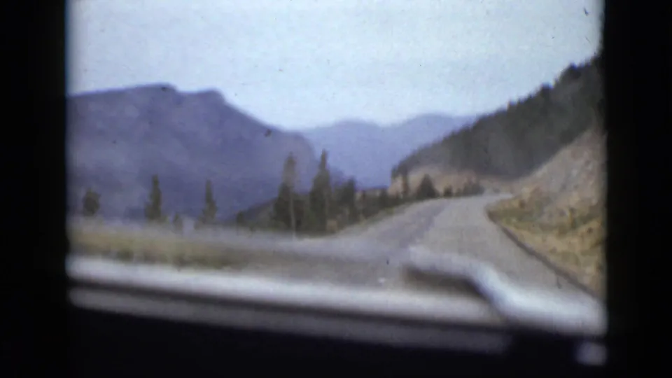 A winding Colorado mountain road recedes through slopes and evergreens, filmed from a moving car with visible windshield edge and 8mm grain.