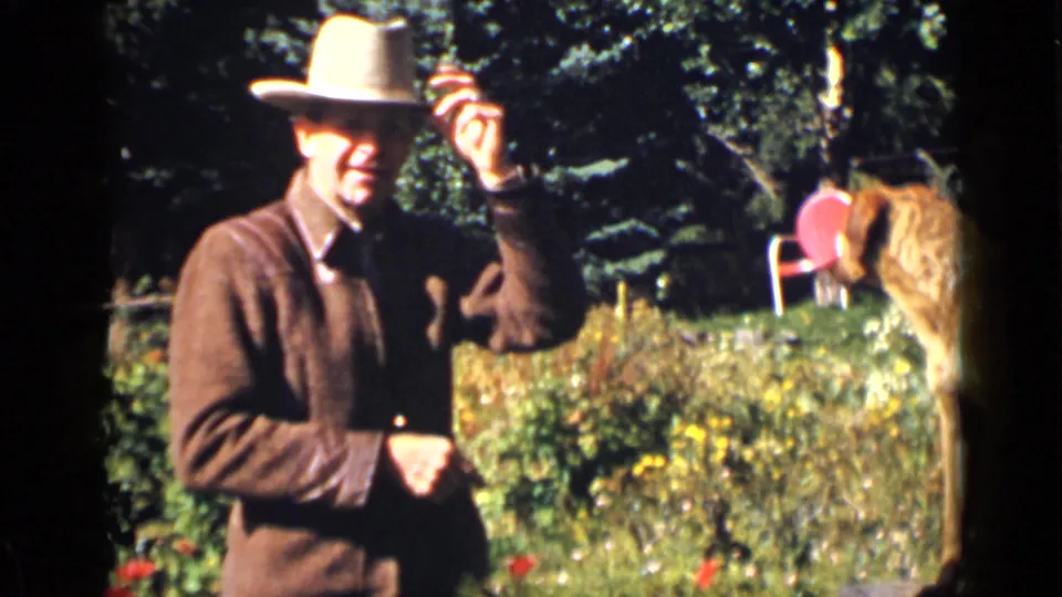 The man tips his light cowboy hat in a Colorado garden as the red dog stands near a red chair.