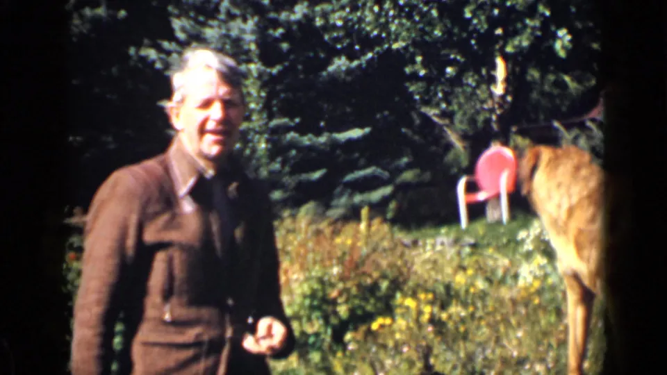 The man appears without his hat in the Colorado garden, standing near the red dog, red chair, trees, and flowers.