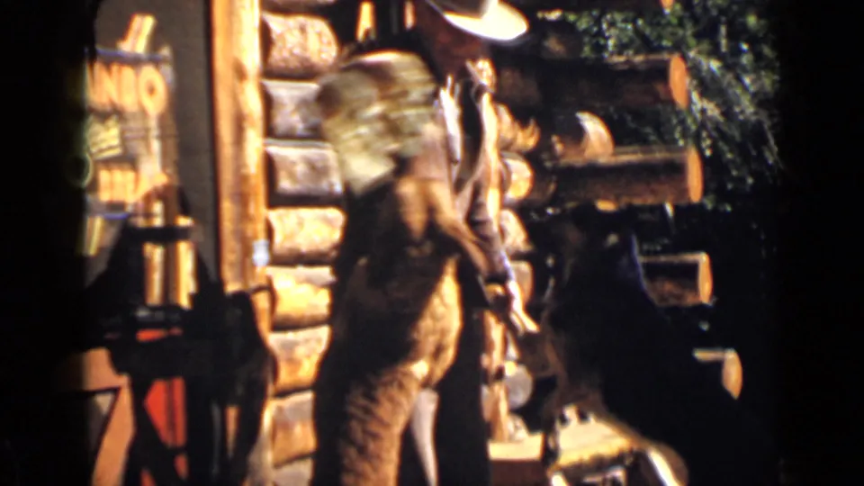 A brown dog rises on its hind legs near a man in a cowboy hat during a playful outdoor moment by a Colorado log cabin.
