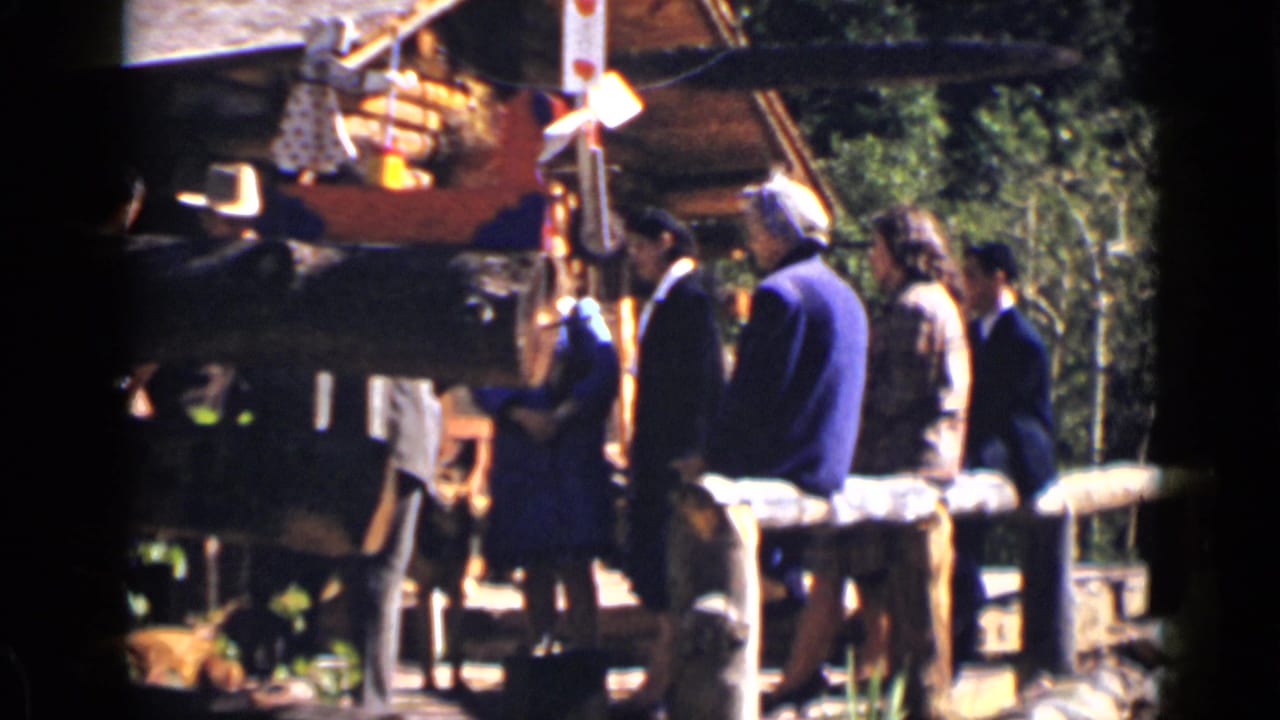 Adults stand near a log ranch structure and wooden fence in bright sunlight.