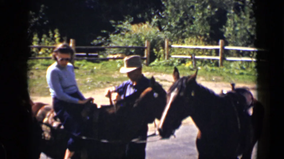 At a Colorado ranch, a woman in sunglasses rides a dark horse as a hat-wearing adult guides the animal.