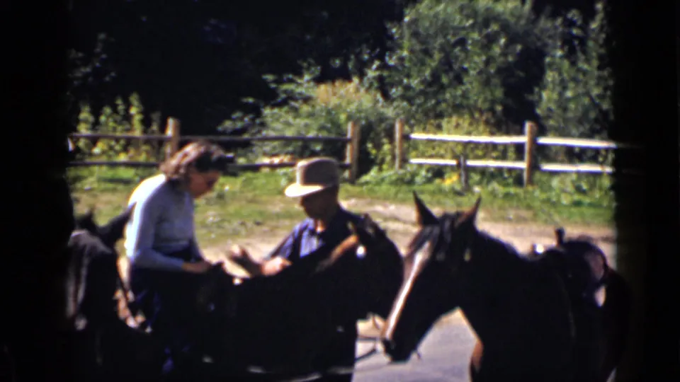The woman rider looks toward her saddle or hands as a man on horseback waits beside her at the ranch.