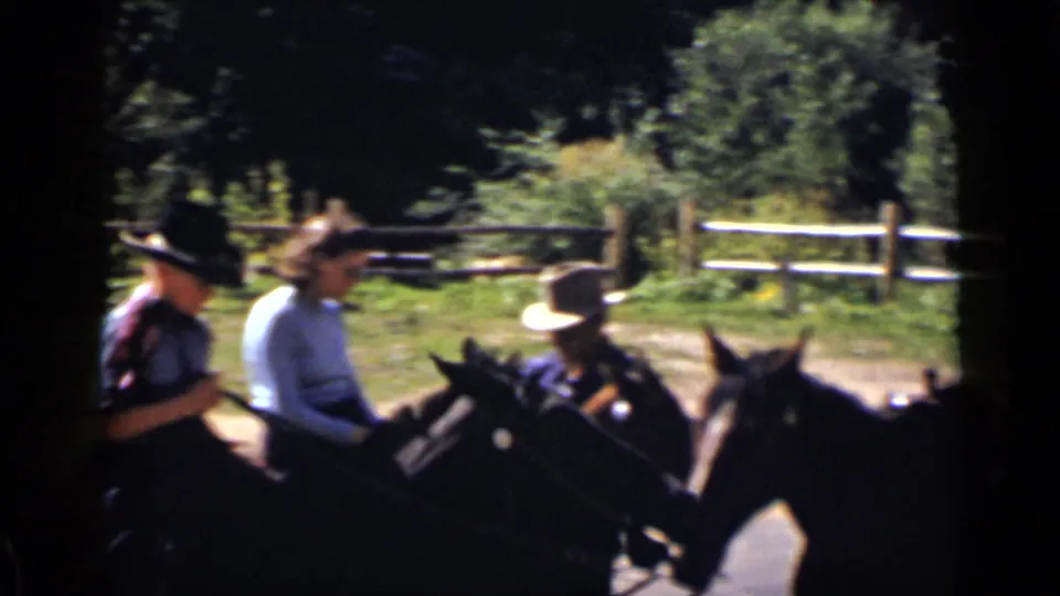 1939 Colorado home-movie frame showing three adults on horseback, with hats, saddles, grass, trees, and a wooden fence nearby.