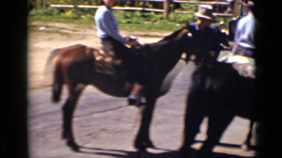 1939 Colorado frame with mounted riders and dark horses on a sunlit paved path, with fencing and greenery in the background.