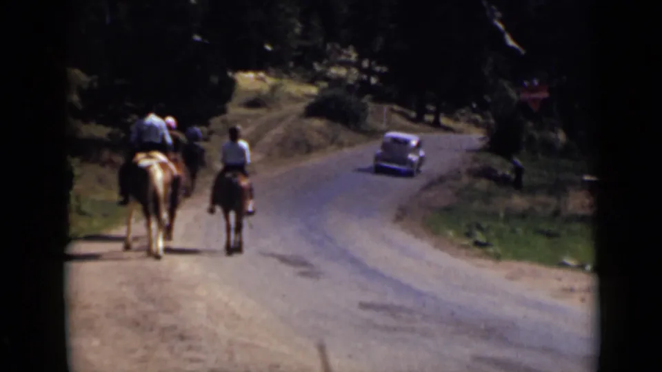 Colorado 1939 frame showing horseback riders from behind on a rural road with a vintage car in the distance near trees.