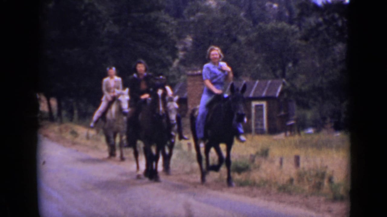 Three people ride horses along a rural road with trees and a small building behind them.