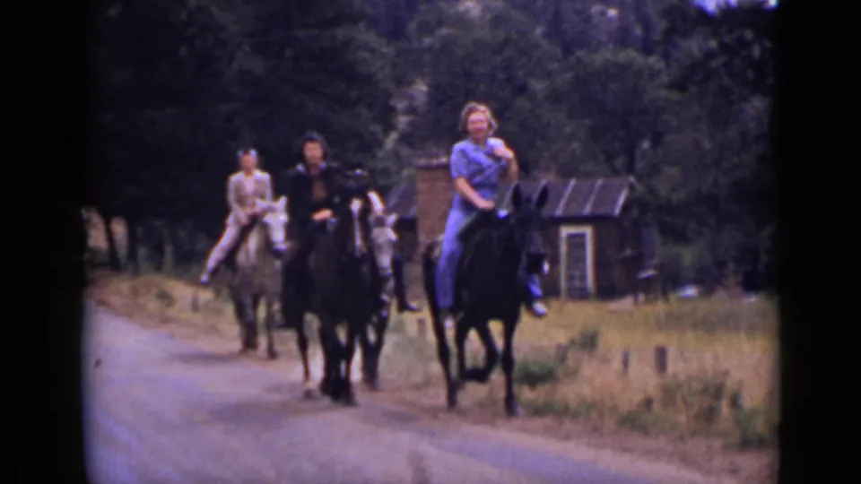 1939 Colorado home-movie frame showing three riders on horseback moving along a rural road near trees and a small rustic structure.