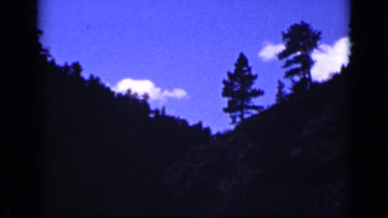 Dark mountain ridge with evergreen trees silhouetted against a blue sky and white clouds.