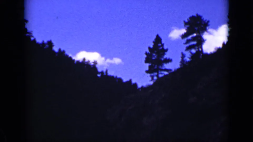 1939 Colorado home-movie frame showing shadowed mountain slopes, ridge trees, blue sky, and small white clouds.
