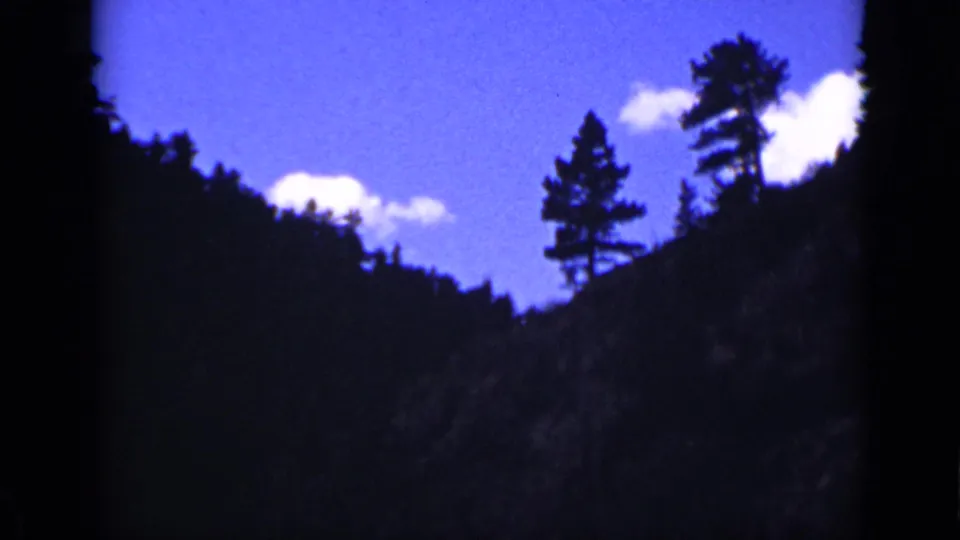 1939 Colorado landscape frame showing a dark valley shape, conifer silhouettes, and bright cloud patches in blue sky.