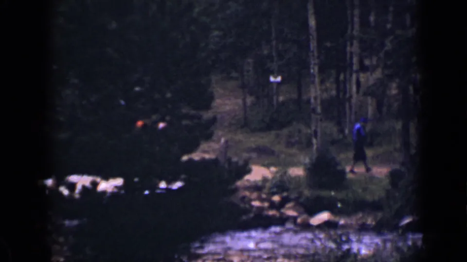 1939 Colorado 8mm frame with a distant figure in blue near a rocky stream or path, surrounded by dense trees and shadows.