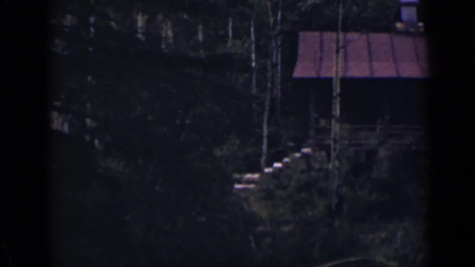 1939 Colorado home-movie frame with a rustic cabin, red roof, porch area, and pale stone steps in a shadowed wooded setting.