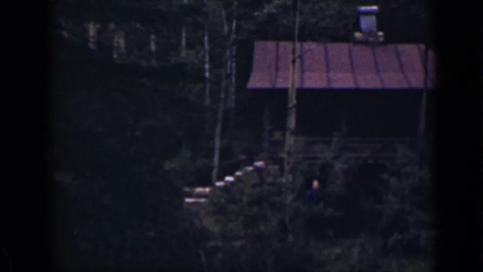 1939 Colorado frame showing a distant person in blue near the stone steps leading up to a wooded cabin with a red roof.