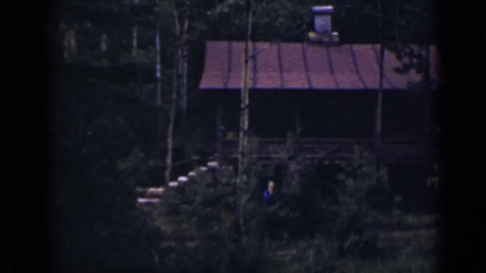 1939 Colorado home-movie frame with a distant person in blue beside a wooded cabin, stone steps, porch railing, red roof, and chimney.