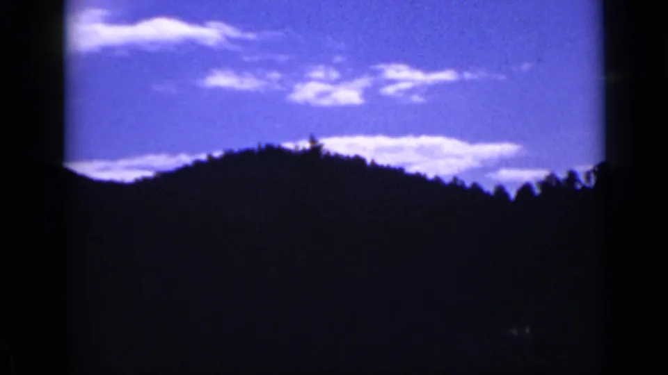 1939 Colorado mountain view with a dark wooded hill, bright blue sky, and scattered clouds in a home-movie frame.