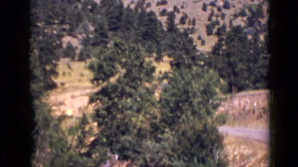 1939 Colorado home-movie frame with a paved curve, stone roadside wall, trees, and scrubby mountain slope.