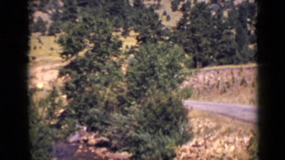 1939 Colorado 8mm frame showing creek water in the foreground below shrubs, a curved road, and wooded slope.