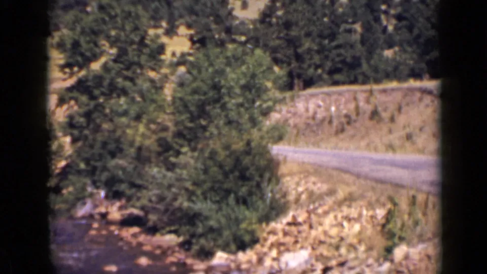 1939 Colorado home-movie frame with creek water, roadside brush, a pale road curve, and pine-covered hillside.