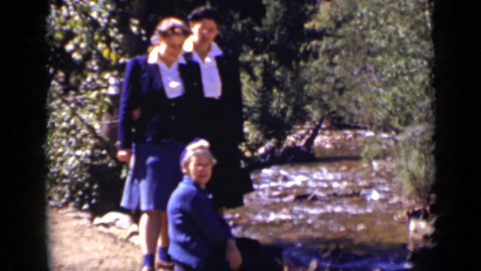Three women pose near a rapidly running stream in Colorado, with two younger women standing and an older woman seated in front.