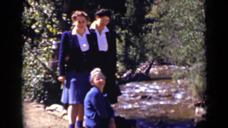 Two younger women in dark sweaters and skirts stand behind a seated older woman beside a wooded Colorado stream.