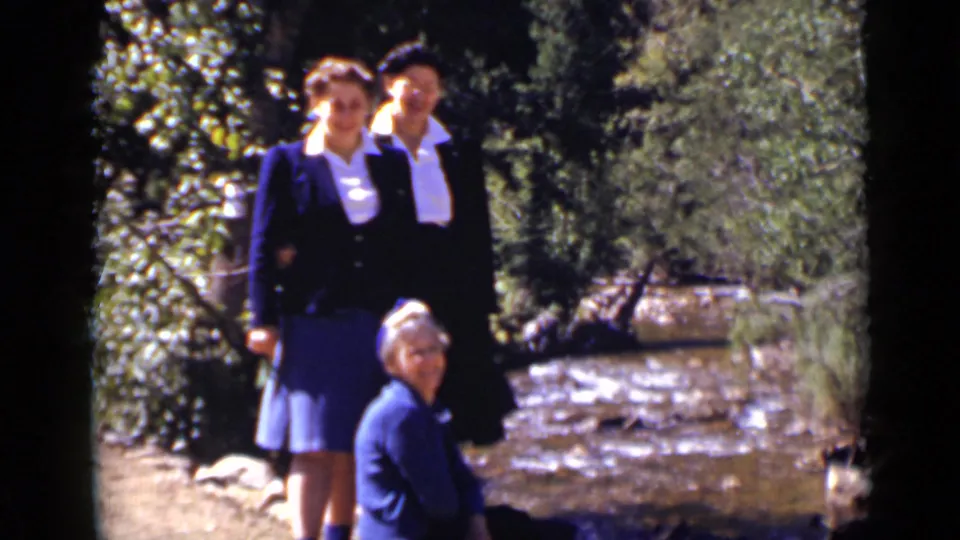 A late-1930s Colorado home-movie frame shows three women by a stream, with wooded banks and sunlit water behind them.