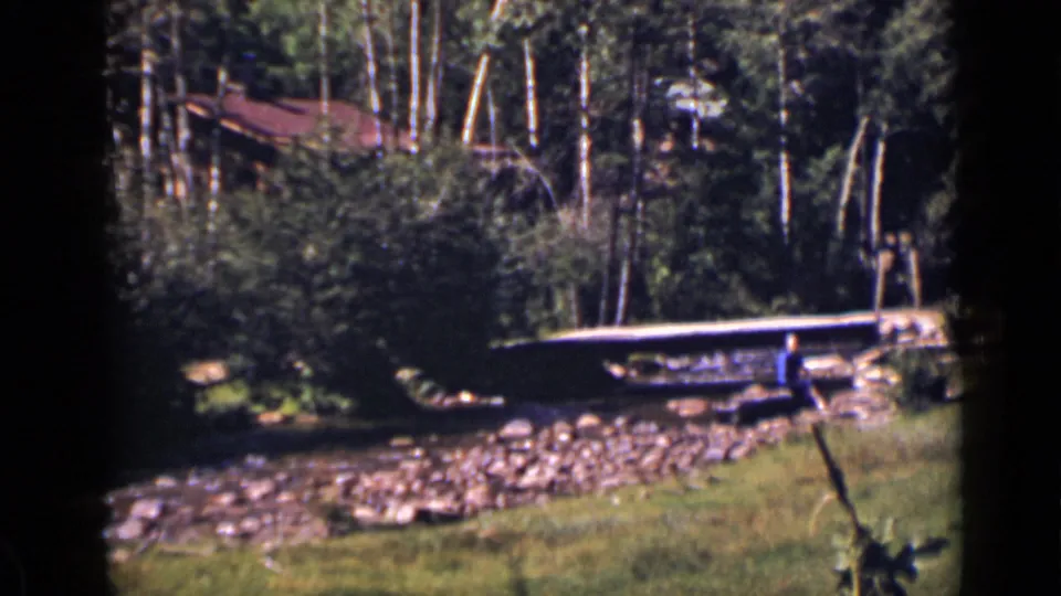 A wooded Colorado creek scene shows a small bridge, rocky bank, and rustic roof in 1939 home-movie footage.