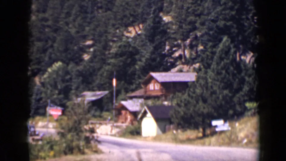 1939 Colorado home-movie frame showing cabins among evergreens, a winding road, hillside terrain, and a red stop sign.