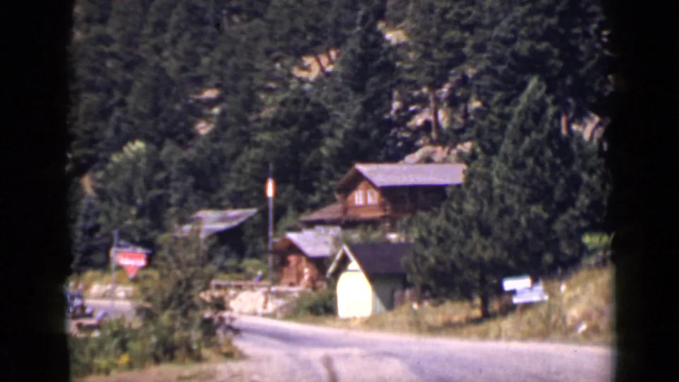 1939 Colorado frame showing a cabin partly screened by evergreens beside a winding road and mountain hillside.