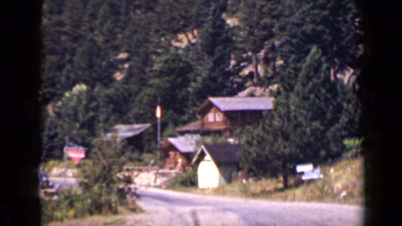 1939 Colorado home-movie frame showing wooden chalets near a mountain road with dense pine forest behind them.