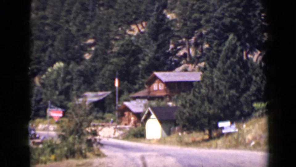 1939 Colorado home-movie frame showing wooden chalets near a mountain road with dense pine forest behind them.