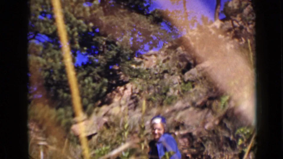 In 1939 Colorado footage, a woman in a blue outfit and head covering stands below rocky hillside vegetation and evergreen trees.
