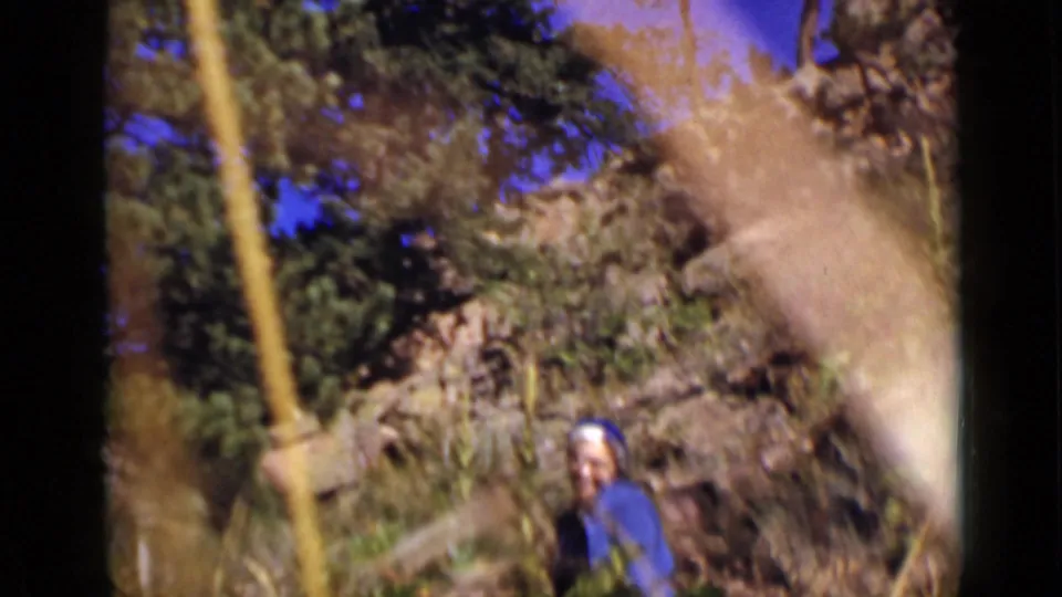 The woman remains on the Colorado hillside, partly small in frame, with trees, brush, rocks, and warm 8mm color shift around her.